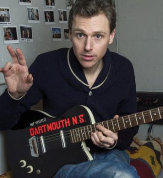 Joel Plaskett holding an electric guitar sitting on a bed pointing at polaroids on the wall beside him